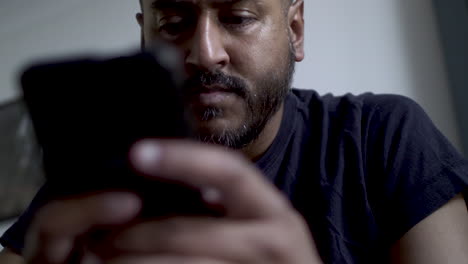 Low-Angle-View-Of-Bearded-Indian-Male-Holding-Smartphone-And-Pensively-Looking