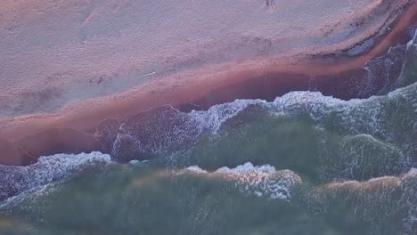 Aerial-birdseye-view-of-the-coastline-of-a-sandy-beach-with-gentle-waves-hitting-the-yellow-sand,-Baltic-sea-in-Liepaja,-Latvia,-golden-hour-light,-wide-angle-point-of-view-drone-shot