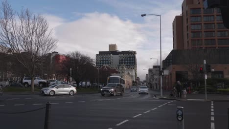 View-up-Campbell-Street-in-Hobart,-as-cars-drive-down-wet-road-with-hospital-in-background,-pedestrians-wait-at-lights-to-cross-road-on-clear-winter-day