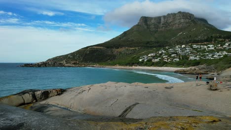 waves-crashing-over-large-boulders-at-Llandudno-Beach-and-Table-Mountain-in-background,-aerial