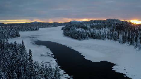 Beautiful-aerial-view-orbiting-right-across-snowy-Lapland-Sweden-winter-lake-landscape-forest-trees-at-sunrise