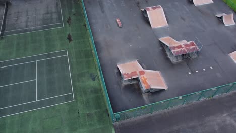 Aerial-view-flying-above-fenced-skate-park-ramp-and-tennis-court-in-empty-closed-playground