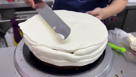 Pastry-chef-using-spatula-to-frost-a-smooth-cake-with-buttercream-meringue-frosting-the-whole-cake-while-spinning-the-revolving-cake-turntable-stand,-close-up-shot-in-commercial-bakery-setting