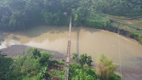 aerial-drone-view-of-suspension-bridge-over-the-river-with-motorcycle-crossing-on-it-in-the-foggy-morning-with-sun-ray,-central-java,-Indonesia