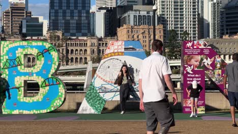People-strolling-by-river-bank-with-tourists-having-fun-taking-photos-with-the-iconic-landmark,-block-sign-of-Brisbane-city-on-a-sunny-day,-Queensland,-Australia,-close-up-handheld-motion-shot
