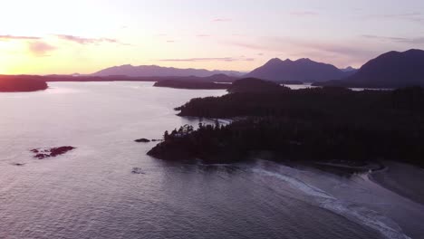 Ein-Schwenkendes-Drohnenvideo-über-Dem-Meer-Neben-Einer-Bewaldeten-Küste-Bei-Sonnenuntergang-Mit-Sichtbaren-Bergen-Im-Hintergrund
