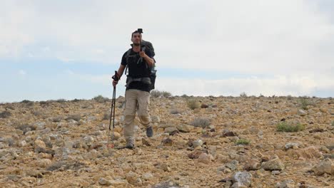 Low-angle-shot-of-a-hiker-talking-on-a-live-video-while-walking-on-Mount-Ramon-desert-trail-on-the-way-to-Ramon-Crater-in-the-Negev-Desert,-Israel-at-daytime