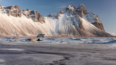 Drone-flight-tracking-shot-of-white-van-arriving-massive-Vestrahorn-Mountain-on-Iceland-during-sunny-day---Snow-covered-rocks-and-landscape-during-cold-winter