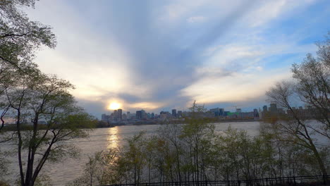 TimeLapse-Montreal-at-Beautiful-Golden-Sunset,-Waterfront-Sightseeing-View-of-St-Lawrence-River,-Water-Flow,-Trees,-Cloudy-Sky-and-Downtown-Buildings-in-Background