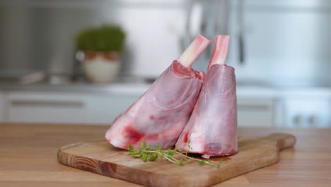Raw-Lamb-Shank-On-Wooden-Chopping-Board-With-Herbs-On-The-Side---close-up,-selective-focus