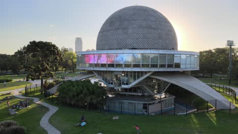 Aerial-dolly-out-of-Galileo-Galilei-Planetarium-and-people-resting-in-Palermo-Woods-at-sunset,-Buenos-Aires