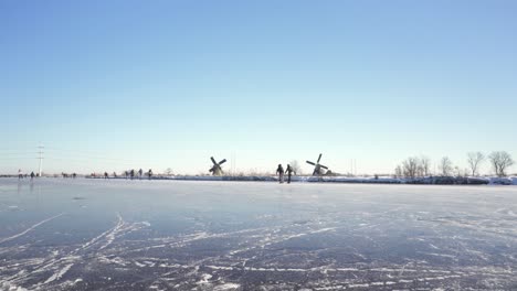 People-ice-skating-on-frozen-Dutch-canal,-windmills-in-background,-winter-scene