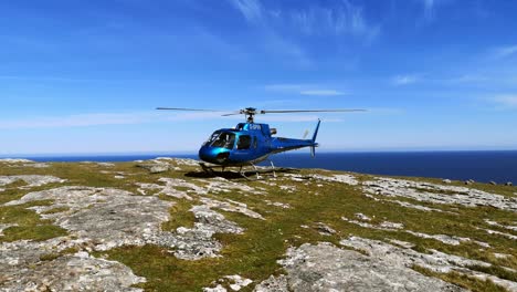 Private-helicopter-perched-on-mountain-cliff-summit-overlooking-blue-ocean-landscape-with-propeller-spin-wide-shot