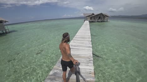 A-young,-fit-and-strong-man-with-long-hair-and-beard-is-walking-on-the-wooden-jetty-during-a-sunny-day-and-smiling-to-the-camera-as-he-film-himself-walking-to-his-luxury-bungalow-from-the-beach