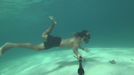 A-young,-fit-and-strong-man-with-long-hair-and-beard-is-swimming-with-snorkeling-goggles-dives-down-from-the-surface-and-explores-the-beautiful-coral-reef-below-with-white-sand-beach-at-the-bottom
