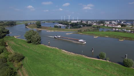 Backwards-aerial-movement-following-an-empty-cargo-vessel-passing-by-on-river-IJssel-near-Zutphen-with-floodplains-and-wind-turbines-in-the-background