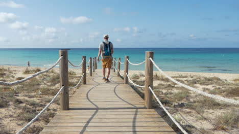 Man-tourist-walking-alone-on-a-wooden-bridge-leading-to-a-golden-sandy-beach-in-Menorca