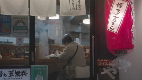 Anciana-Comiendo-Sola-En-Un-Restaurante-Con-Gente-Pasando-Durante-La-Noche-En-Tokio,-Japón