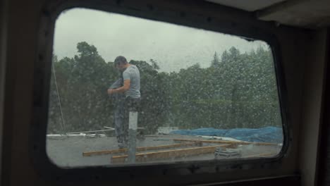 View-through-rainy-boat-window-man-removes-jacket-to-being-working
