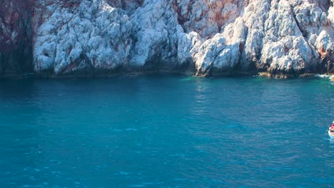 Couple-in-a-white-boat-sailing-near-the-cliffs-at-port-of-Alanya-in-sunny-summer-day,-blue-sea,-handheld-medium-shot-from-a-boat