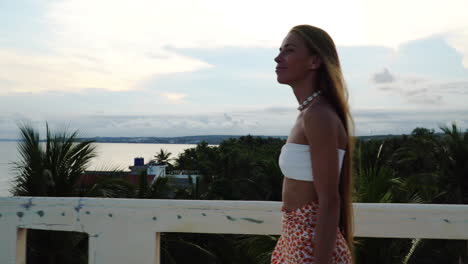 Young-girl-in-white-crop-top-walks-on-a-balcony-with-beach-at-far-end-at-sunset