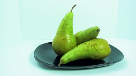 Fresh-big-green-pears-rotates-slowly-on-a-black-plate-on-light-blue-background,-healthy-food-concept,-medium-shot,-camera-rotate-left