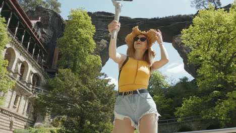 Young-Woman-in-Bohemian-Switzerland-National-Park,-Czech-Republic,-Under-Natural-Arch-and-Old-Hotel-Recording-Selfie-Video-With-Smartphone-and-Gimbal,-Slow-Motion