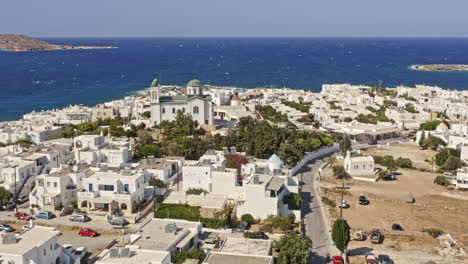 Naousa-Paros-Greece-Aerial-v2-low-flyover-small-village-capturing-faneromeni-church-surrounded-by-beautiful-coastal-whitewashed-buildings-with-paros-park-hilltop-in-the-background---September-2021