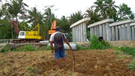 Lonely-farmer-from-Indonesia-hoeing-soil-with-massive-industrial-machines-in-backgorund
