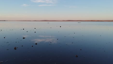 Flying-low-over-the-pink-lake-and-Lochiel-in-South-Australia,-with-rocks-on-the-edge-of-the-water-leading-to-clouds-reflecting-off-the-still-water