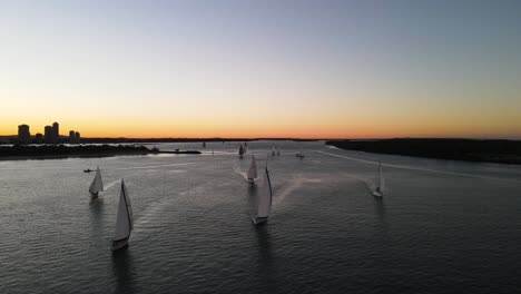 A-group-of-moving-sailing-boats-racing-along-the-calm-waters-of-a-city-harbor-against-a-colorful-sunset-backdrop