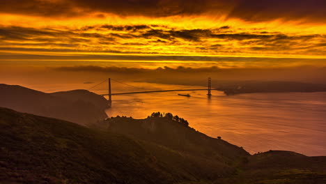 5K-Time-lapse-shot-of-Golden-Gate-Bridge-during-beautiful-yellow-sunrise-and-boat-cruising-below-bridge---Epic-lighting-of-sky-with-flying-clouds---aerial-wide-shot