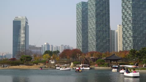 Mucha-Gente-Navegando-En-Pequeños-Botes-De-Viaje-En-Un-Lago-En-El-Parque-Central-De-Songdo-En-Otoño-Con-Modernos-Rascacielos-Altos-En-El-Fondo---Ciudad-De-Incheon,-Corea