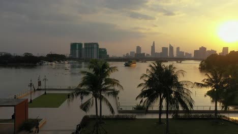 Beautiful-tropical-sunset-park-scene-from-drone-with-people-enjoying-leisure-activities-like-skating,-running-and-cycling-and-a-view-to-city-skyline-and-boats-and-river
