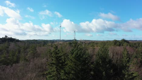 Two-huge-wind-turbines-in-Lindesnes-wind-park-southern-Norway---Aerial-with-parallax-effect-and-spruce-trees-in-foreground---Sunny-clear-day-and-blue-sky---One-turbine-rotating-and-one-stopped