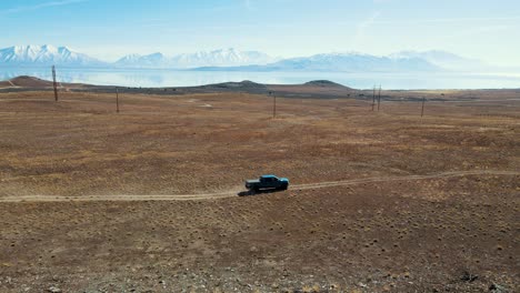 Four-wheel-drive-truck-driving-along-a-dirt-road-with-a-lake-and-snowy-mountains-in-the-distance---aerial-view