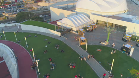 drone-aerial-video-of-outdoor-terrace,-chill-people-sitting-on-green-synthetic-grass,-chilling,-outside-costa-rica-shopping-mall,-drone-video-in-shopping-mall-at-sunset,-outdoors,-outdoor-concert