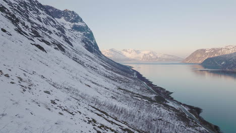 Luftaufnahmen-Entlang-Eines-Steilen,-Schneebedeckten-Berghangs,-Einer-Malerischen-Straße-Neben-Dem-Fjord
