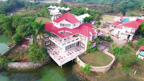 Rotating-aerial-drone-view-of-a-luxurious-holiday-cottage,-to-relax-and-enjoy-swimming-in-the-lake-and-pool,-next-to-the-palm-trees-and-original-vegetation-of-Vadodara,-India