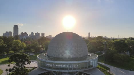 Aerial-pedestal-up,-overlooking-at-beautiful-bright-sun-shinning-behind-famous-building-structure-Galileo-Galilei-Planetarium-in-Palermo-Woods,-downtown-Buenos-Aires