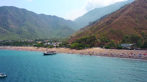 aerial-drone-view-of-Adrasan-beach-full-of-people-as-a-boat-is-anchored-on-the-beach-waiting-for-tourists-on-a-hot-summer-day-in-the-dry-mountain-landscape-of-Turkey