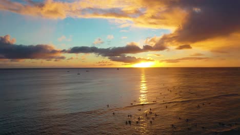 Drone-View,-Radical-Surfers-Catching-Sets-of-Waves-at-Sunset-On-Famous-Waikiki-Beach,-Dawn-Patrol-Hang-Ten-With-Tourists-and-Colorful-Sky-In-Honolulu,-Hawaii