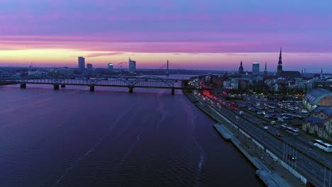 A-spectacular-city-panorama-of-Riga-opens-up-from-the-air:-bridges-across-the-River-Daugava,-intense-traffic,-city-landmarks-are-seen-on-the-sunset