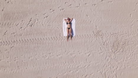 Young-Caucasian-woman-in-bikini-lies-on-white-blanket-on-sandy-beach