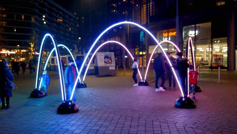 Besucher-Interagieren-Mit-Der-Farbenfrohen-Winterfest-LED-Kunstwerkinstallation-Mit-Beleuchteten-Bögen-Im-Wembley-Park-Bei-Nacht