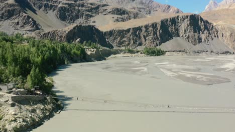 Aerial-drone-flying-backwards-as-tourists-cross-the-famous-Hussaini-bridge-in-Hunza-Pakistan-with-a-fast-river-flowing-below-and-a-green-forest-bordering-the-river