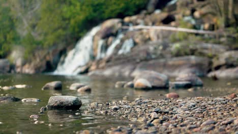 Rocky-river-bank-with-gentle-waves-washing-across-shoreline-with-smooth-stones-and-flowing-waterfall-in-the-background