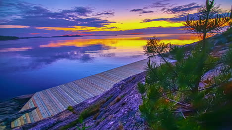 View-of-yellow-sunset-over-blue-sky-beside-blue-lake-next-to-wooden-jettie-on-rocks-in-timelapse-in-the-evening