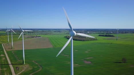 Aerial-view-of-wind-turbines-generating-renewable-energy-in-the-wind-farm,-sunny-spring-day,-low-flyover-over-green-agricultural-cereal-fields,-countryside-roads,-drone-dolly-shot-moving-right