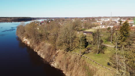 Latvian-rural-landscape-with-a-winding-river-near-Lielvarde-city,-leafless-trees,-aerial-top-view,-Daugava-river,-sunny-spring-day,-wide-revealing-shot-moving-backwards,-camera-tilt-up